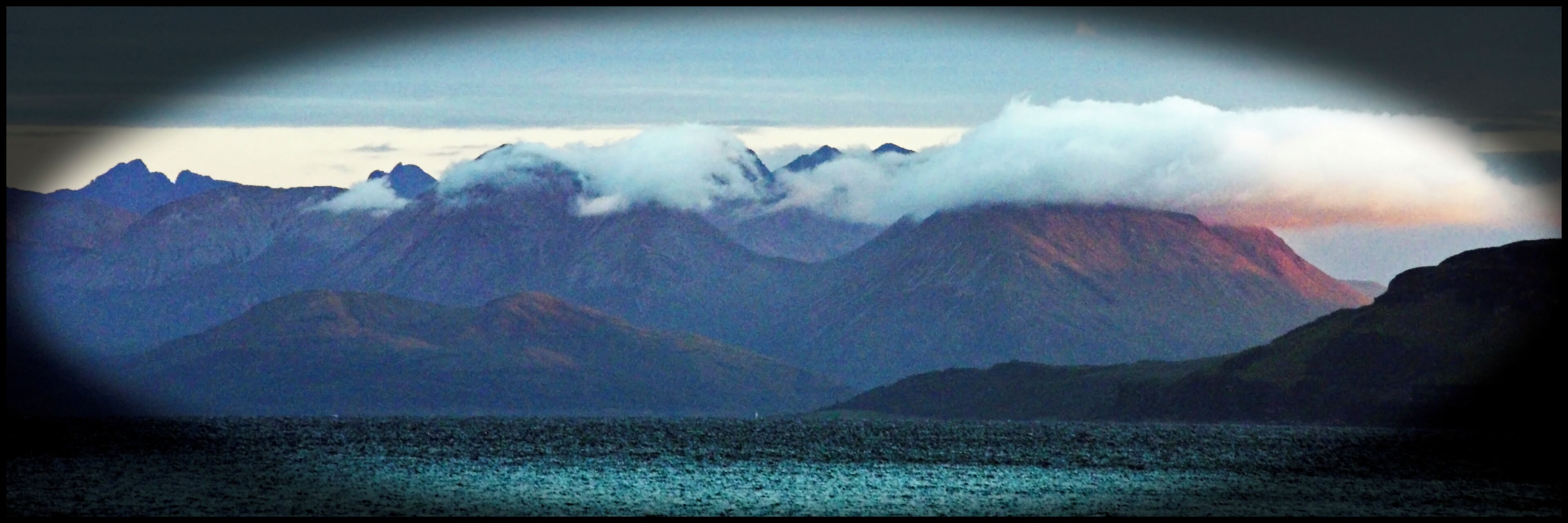 A mountain range with clouds at the peaks and a body of water in the foreground.
