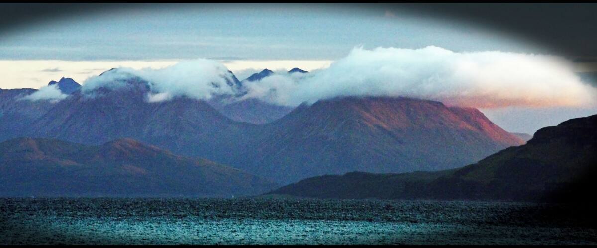 A mountain range with clouds at the peaks and a body of water in the foreground.