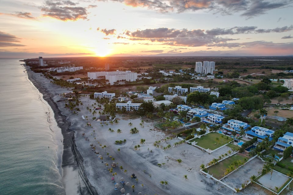A coastal cityscape with a beach, palm trees, and high-rise buildings during sunset.