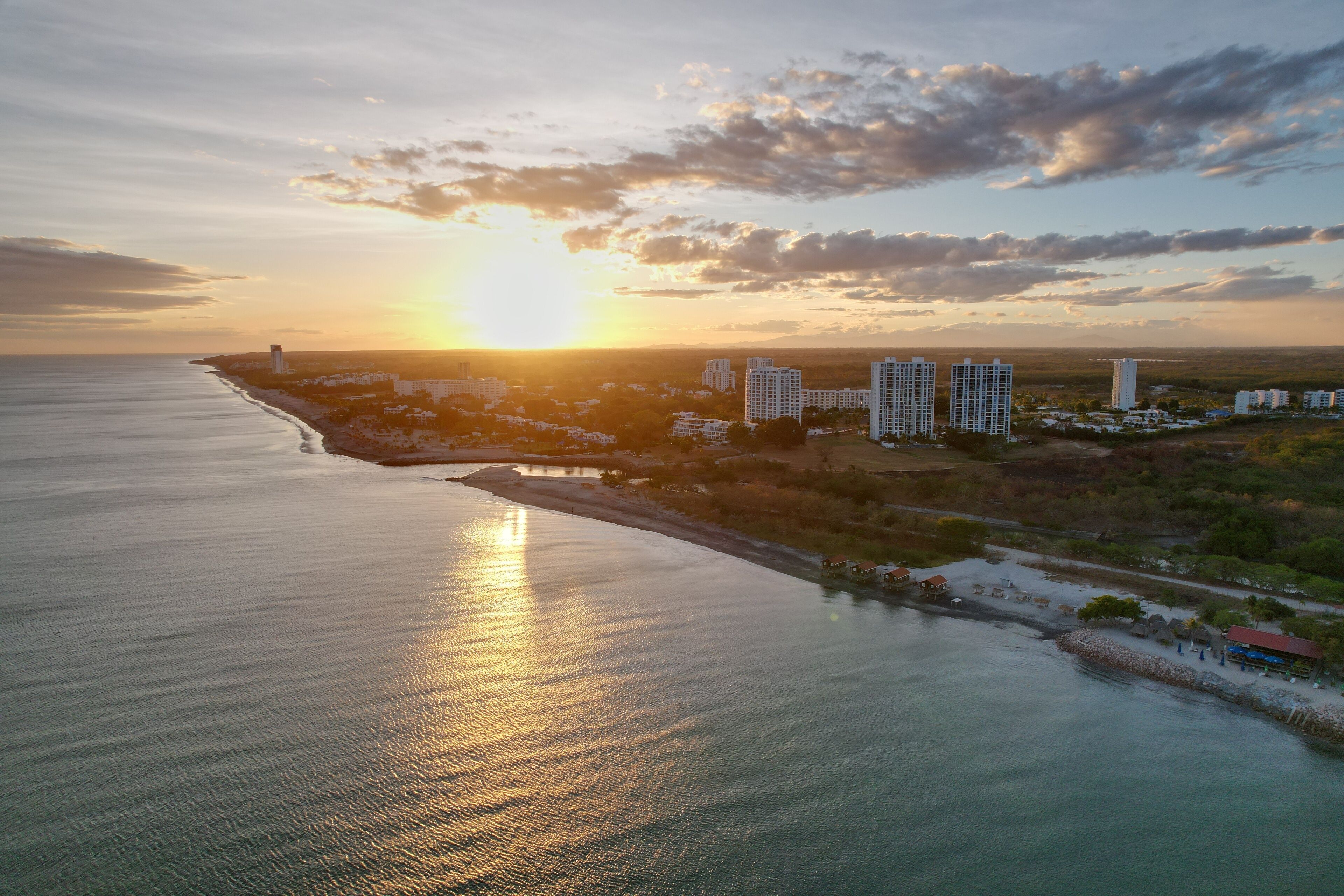 A coastal cityscape with a sunset over the ocean, high-rise buildings, and a beach area.