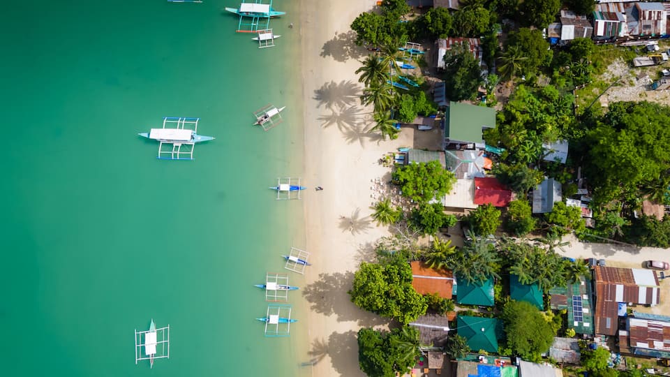 A coastal area with boats, a beach, and buildings surrounded by trees.