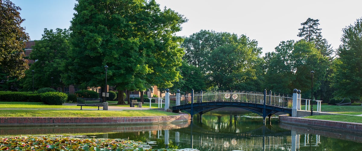 Green trees and pond with reflective surface.  Pedestrian bridge over clear smooth pond.  Central College campus in Pella, Iowa.