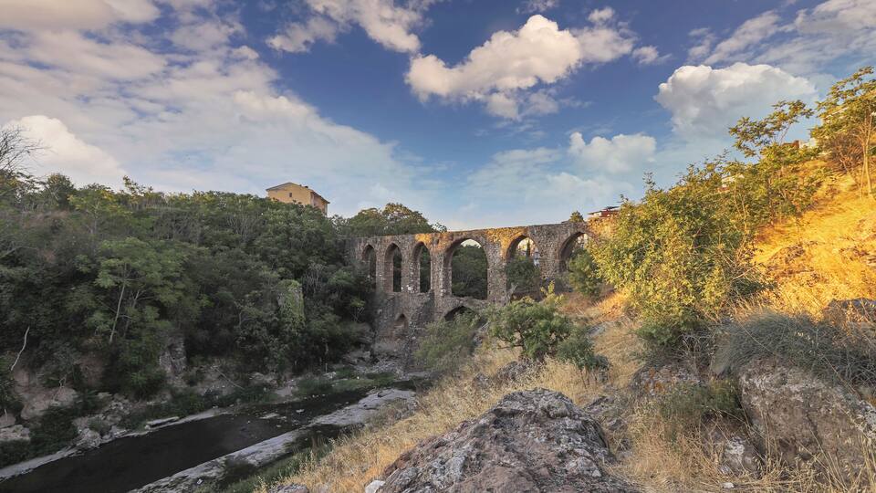 A stone bridge over a river, surrounded by trees and rocky terrain.