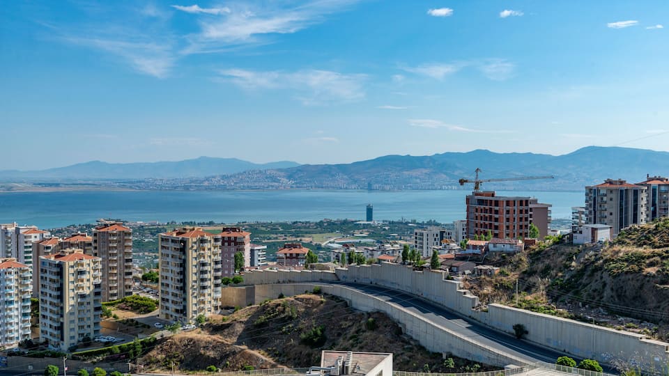 A coastal cityscape with high-rise buildings, a construction crane, and a view of the sea and distant mountains.