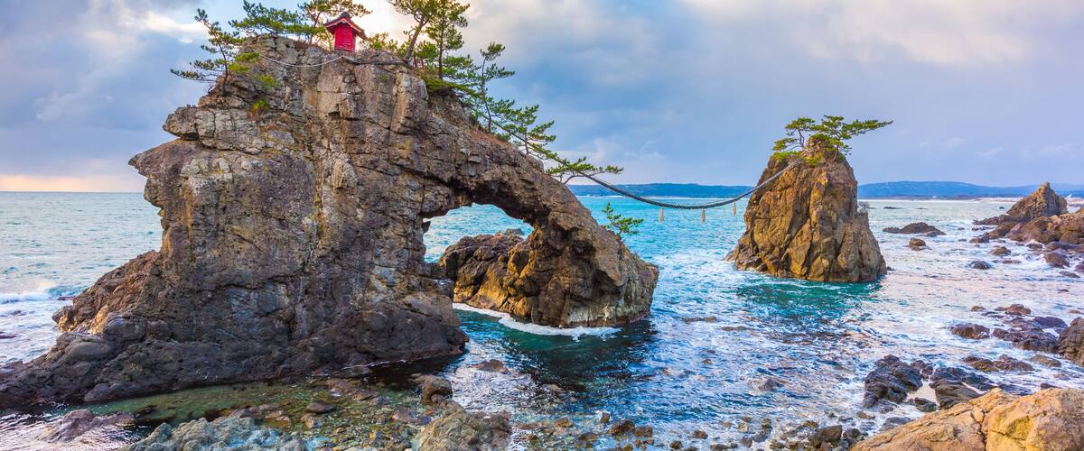 A coastal landscape with a prominent rock arch formation and a red building on top.