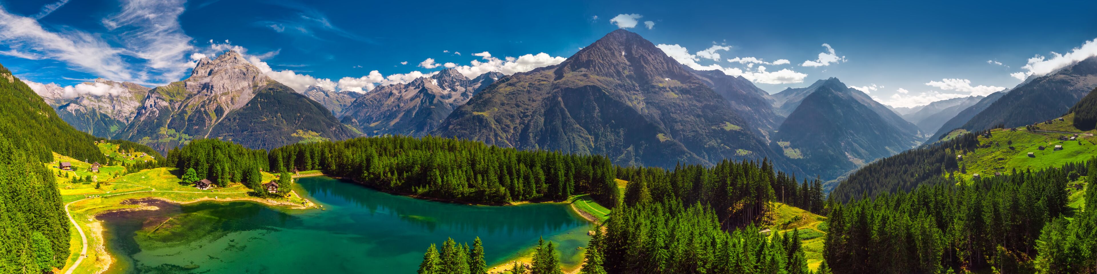 Paisagem montanhosa com um lago azul-turquesa rodeado por florestas densas e um céu azul límpido.