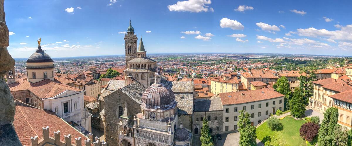 A panoramic view of a historic city with prominent domed and spire-topped buildings, a cathedral, and a distant green landscape.