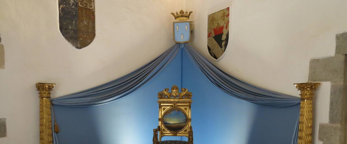 A throne with a crown, flanked by two lion statues, in a room with blue drapes and a shield on the wall.