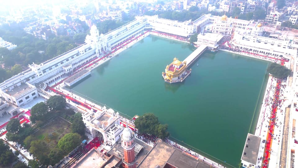 A large square body of water with a golden temple in the center, surrounded by white buildings and a red carpet.