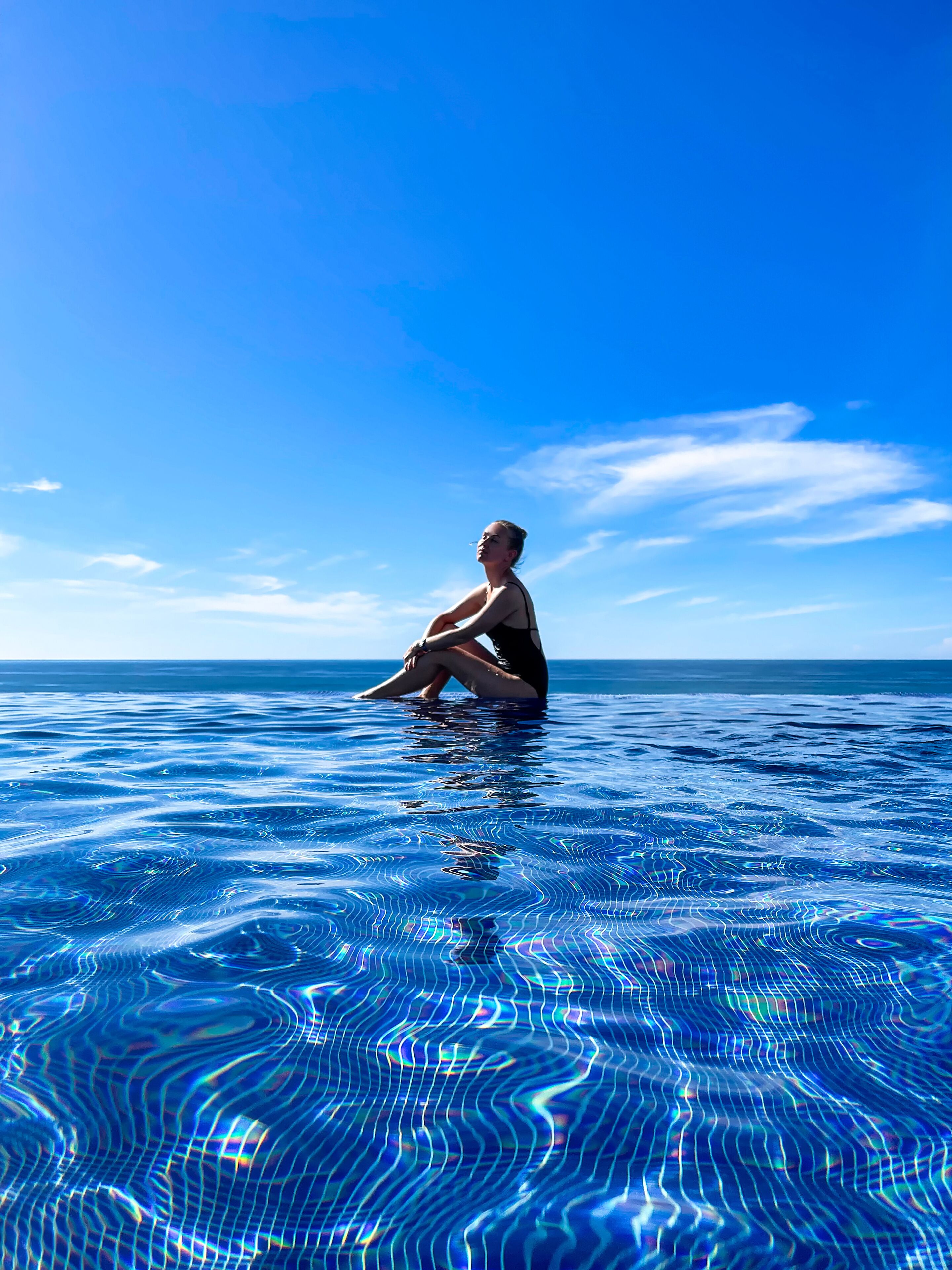 A person sitting in a swimming pool with a clear blue sky and ocean in the background.