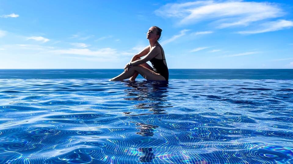 A person sitting in a swimming pool with a clear blue sky and ocean in the background.