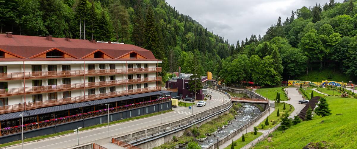 A riverside hotel with a red roof, a road alongside the river, and a forested hillside in the background.