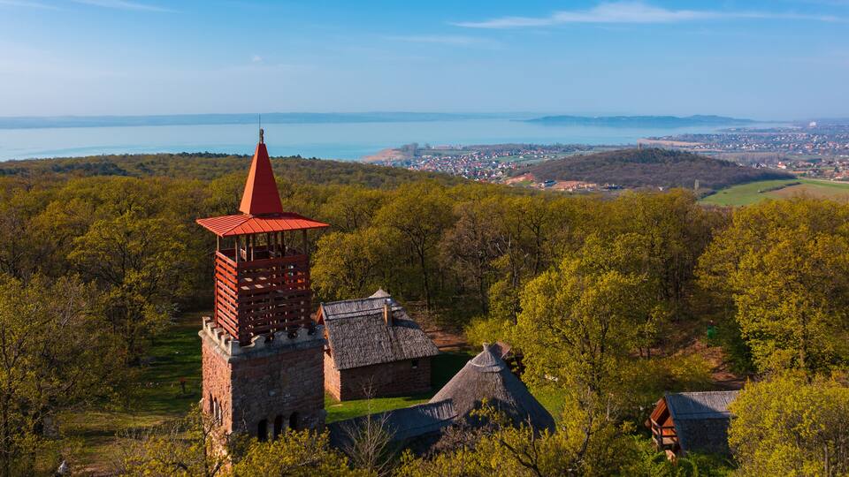 A tower with a red roof, a stone building, and a thatched-roof structure surrounded by trees and a distant town by a body of water.