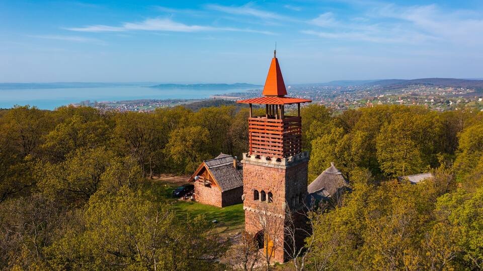 A tower with a red roof and a bell tower in a forested area with a view of a town and a body of water in the distance.