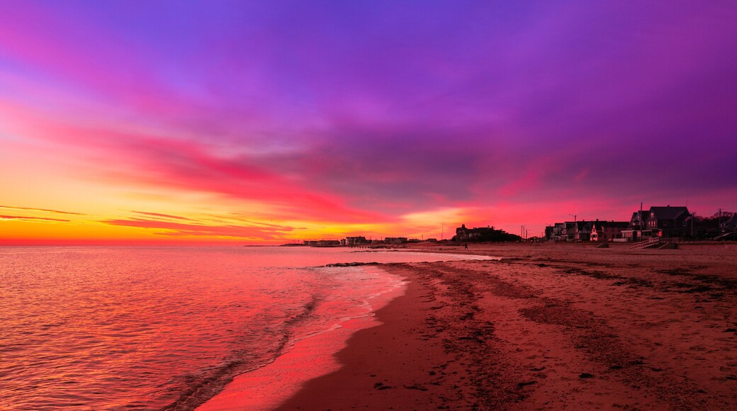 Vibrant saturated pink and blue seascape at sunrise. Curved beach, gentle waves, dramatic clouds over the coastal village. Stunning red twilight landscape over Falmouth Height Beach on Cape Cod.