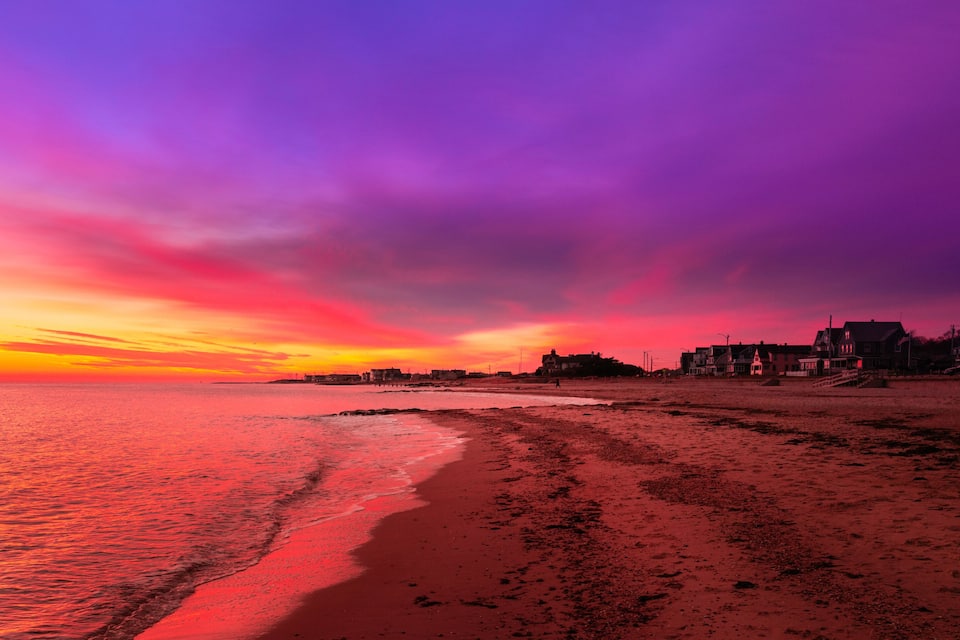 A beach at sunset with a vibrant sky, ocean waves, and buildings along the shore.