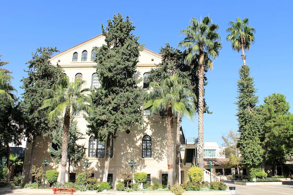A historic building with arched windows and a red roof, surrounded by tall palm trees and well-maintained gardens.