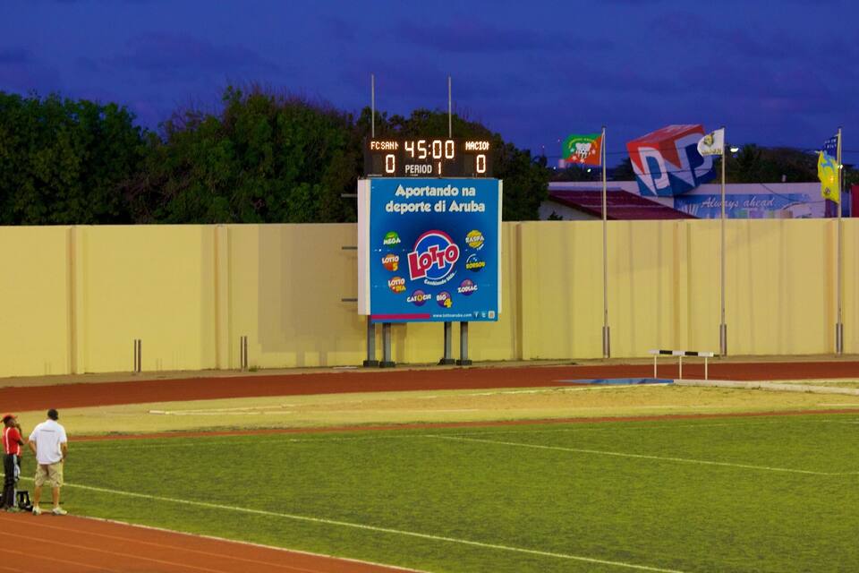 Un camp esportiu amb un marcador, un cartell gran i banderes.