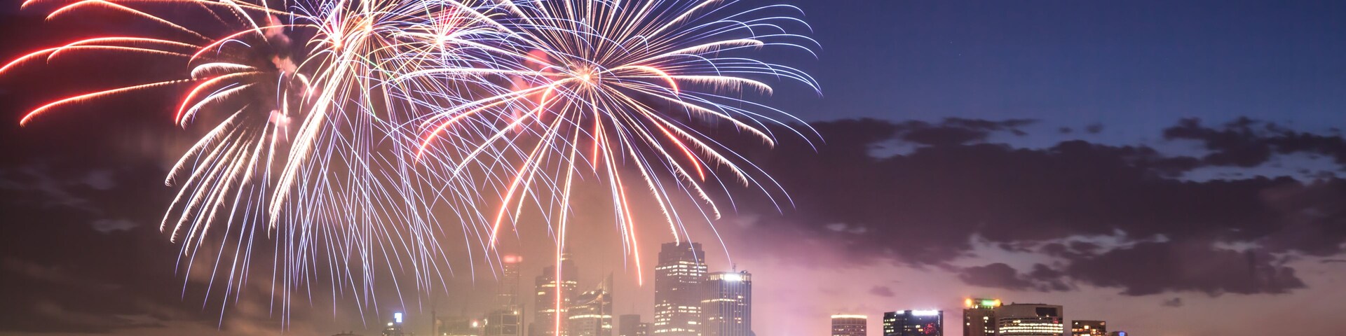 Silhueta urbana com fogos de artifício explodindo sobre a água.