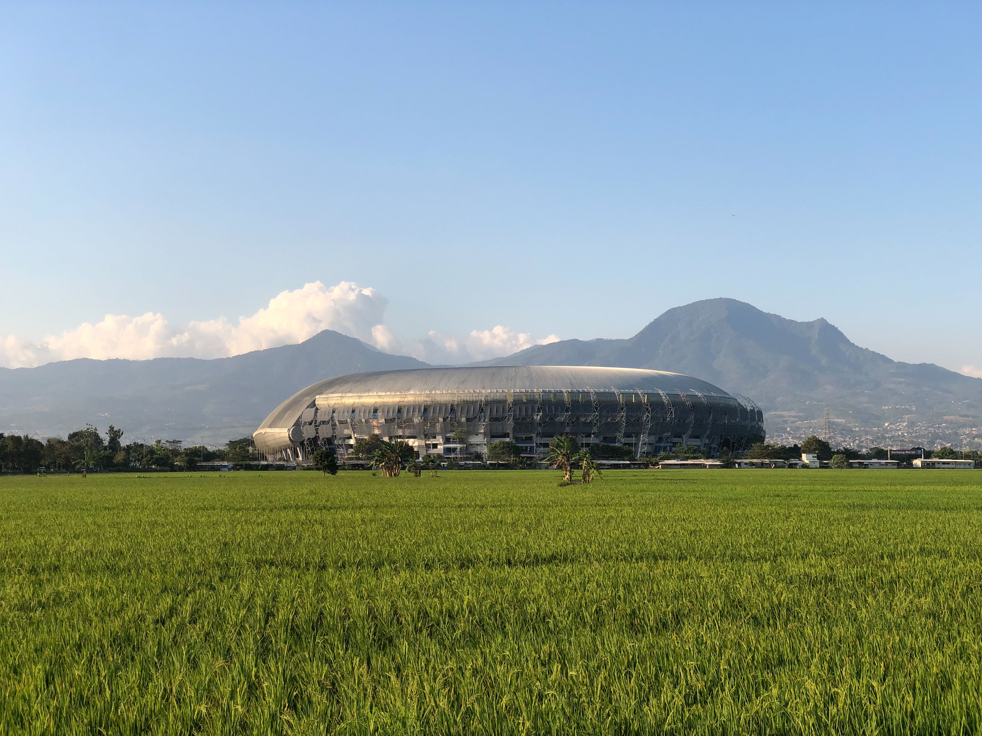 A stadium with a large, curved roof, surrounded by a green field and mountains in the background.