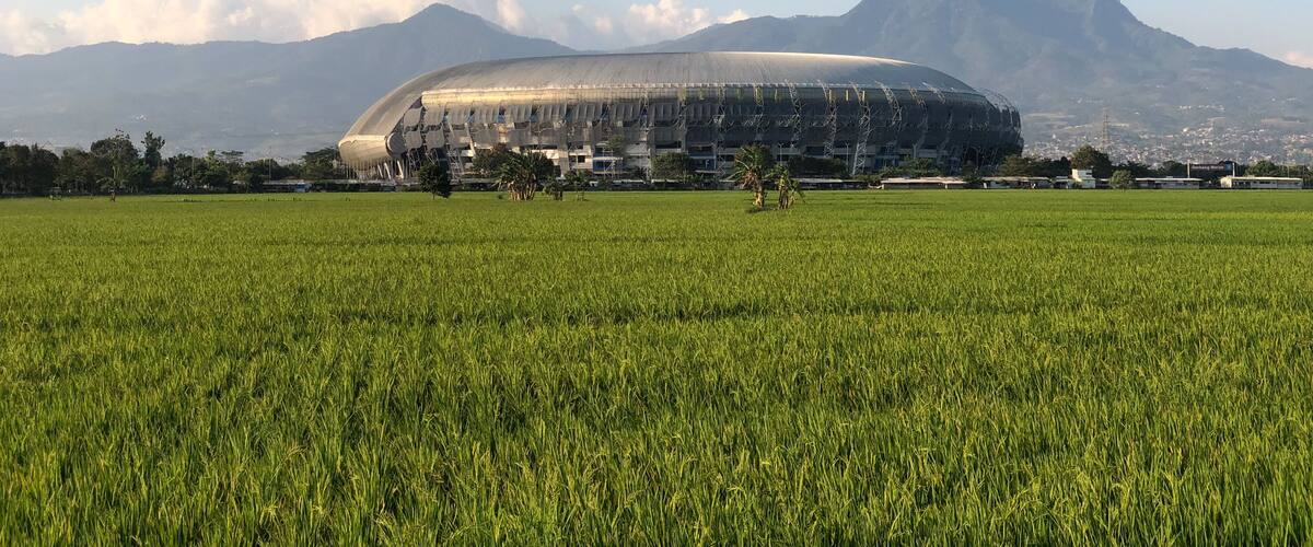 A stadium with a large, curved roof, surrounded by a green field and mountains in the background.