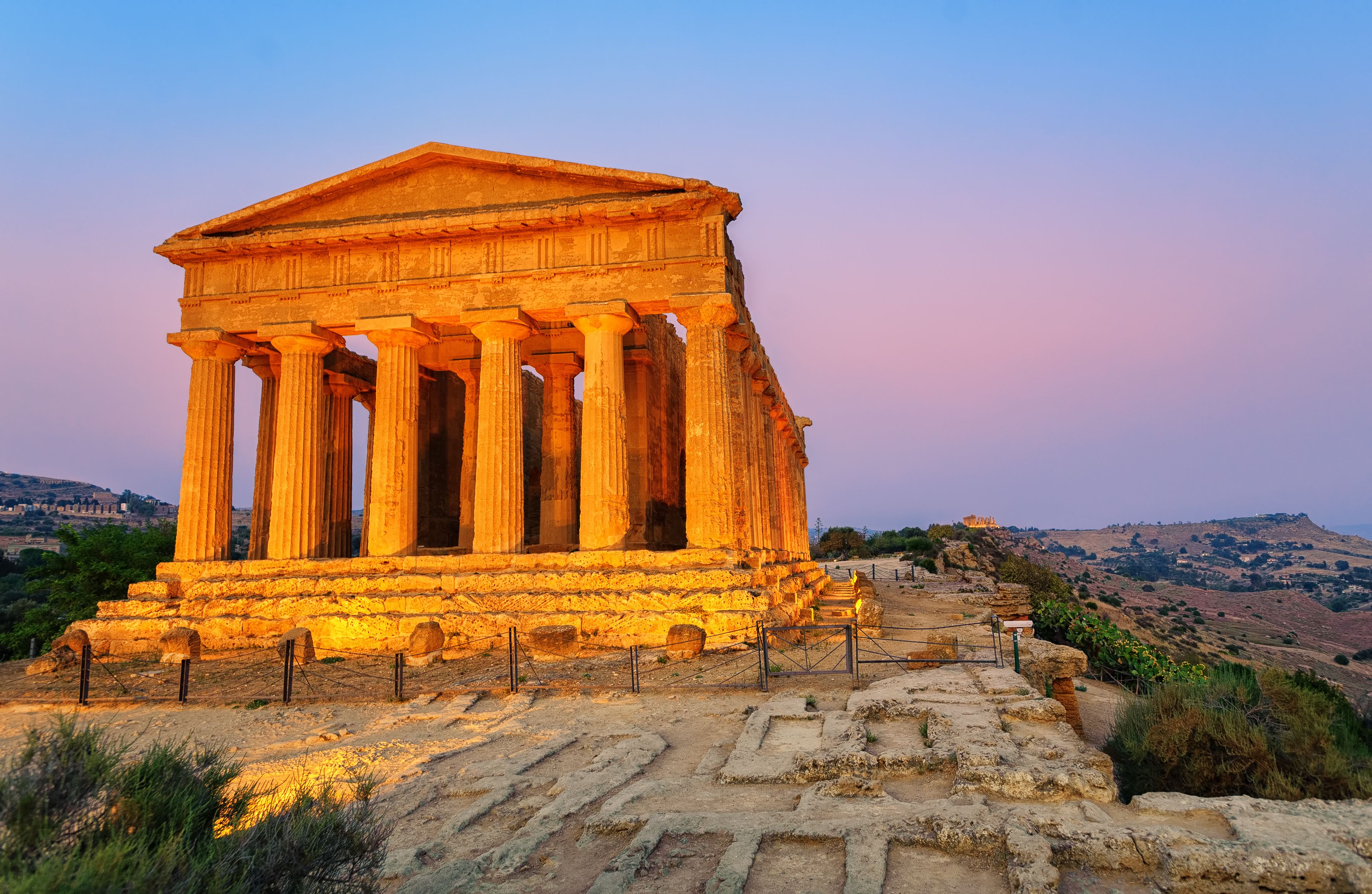 An ancient Greek temple with columns and a pediment, set against a hilly landscape during sunset.