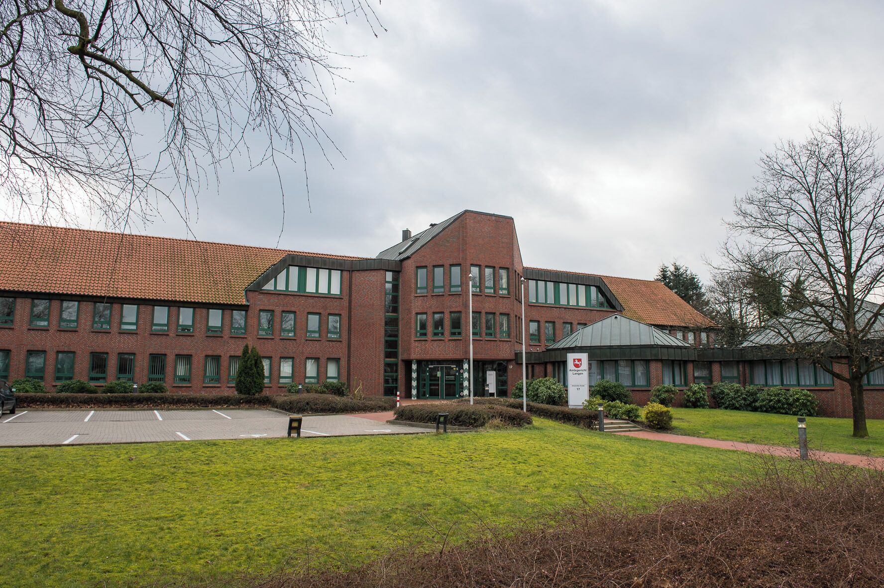 A large red-brick building with green windows and a tiled roof, surrounded by a grassy area and a few trees.