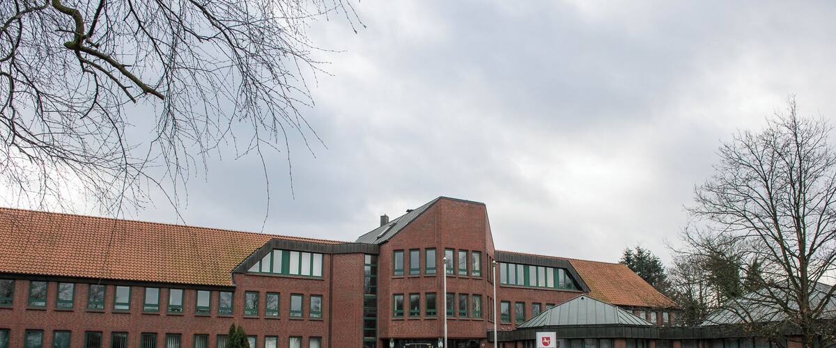 A large red-brick building with green windows and a tiled roof, surrounded by a grassy area and a few trees.