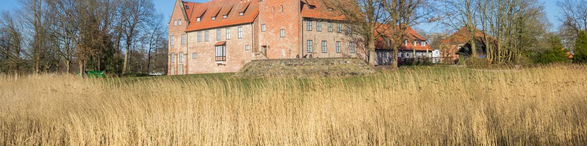 A historic brick building with a red-tiled roof, situated by a calm body of water with tall reeds in the foreground.