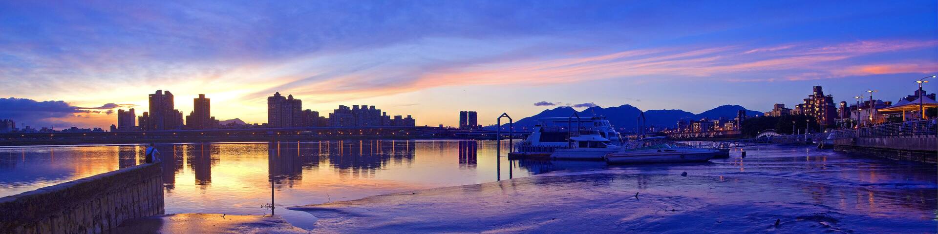 Un paisaje urbano frente al río al atardecer, con aguas tranquilas que reflejan el cielo.