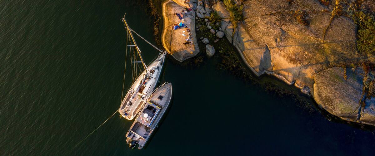 A sailboat anchored near a rocky coastline with a small group of people onshore.