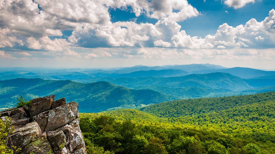 A mountainous landscape with dense green forests and a clear blue sky with clouds.