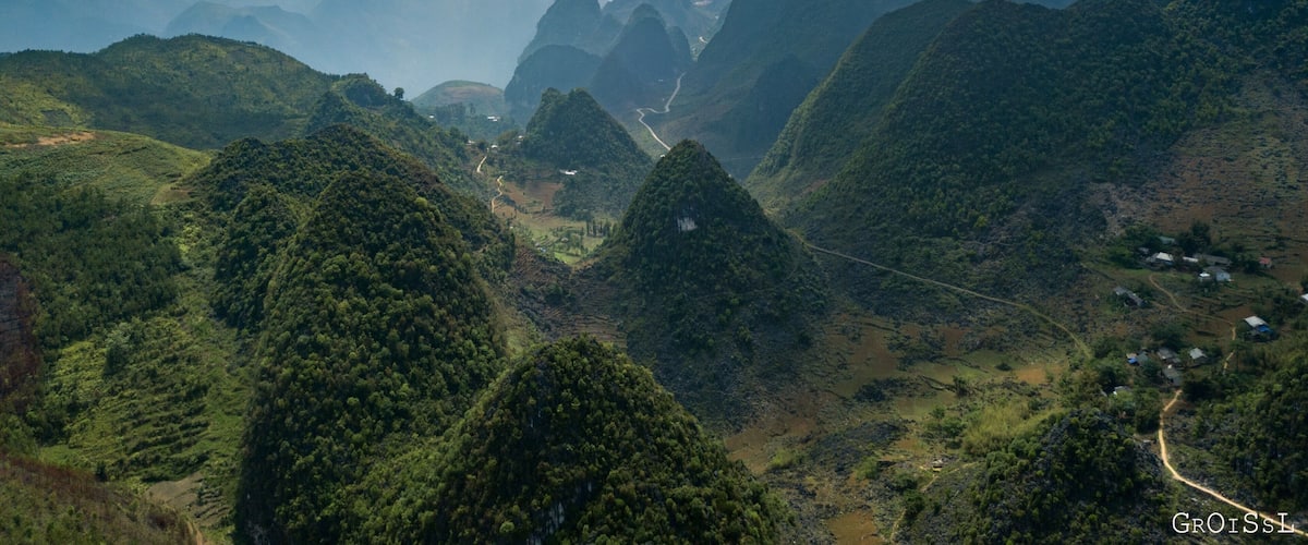 A mountainous landscape with lush greenery, winding roads, and distant peaks under a cloudy sky.