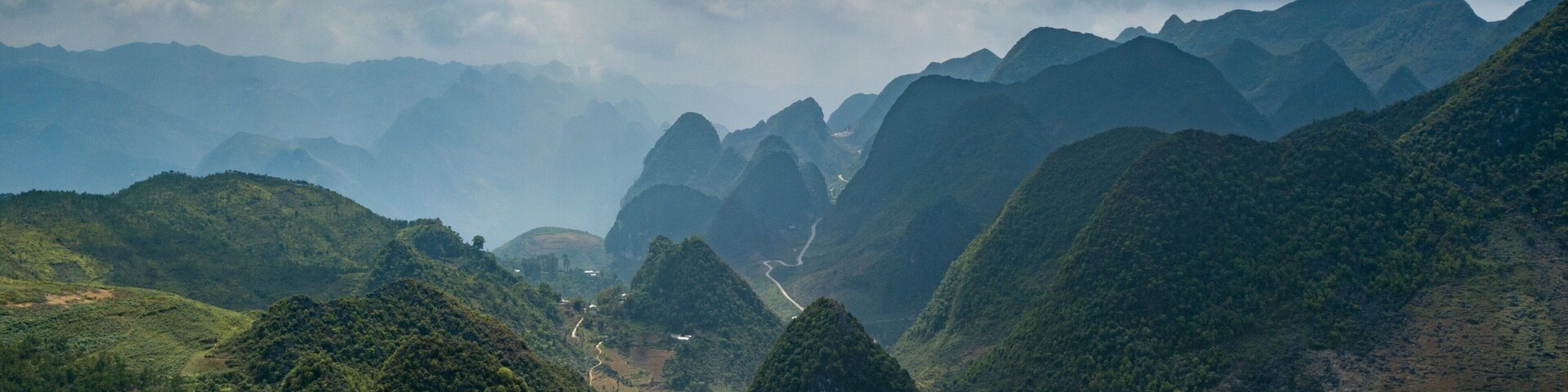 A mountainous landscape with lush greenery, winding roads, and distant peaks under a cloudy sky.