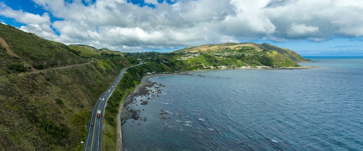 Coastal highway winds through New Zealand's scenic landscape, hugging the coastline. Traffic moves along the road, nestled between hills and the ocean. PUKERUA BAY, PORIRUA, WELLINGTON, NEW ZEALAND