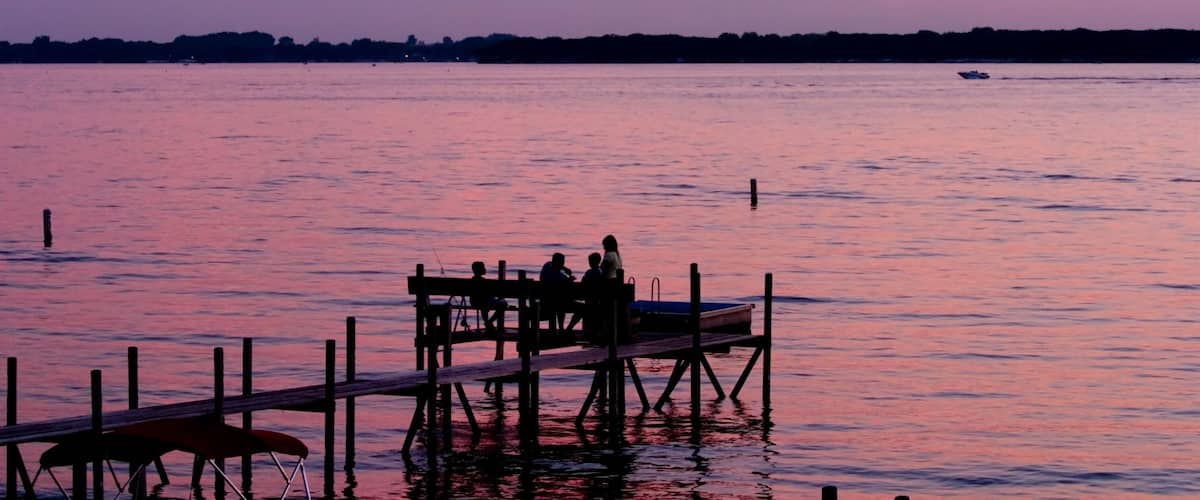 A sunset over a calm lake with a dock and people sitting on it.