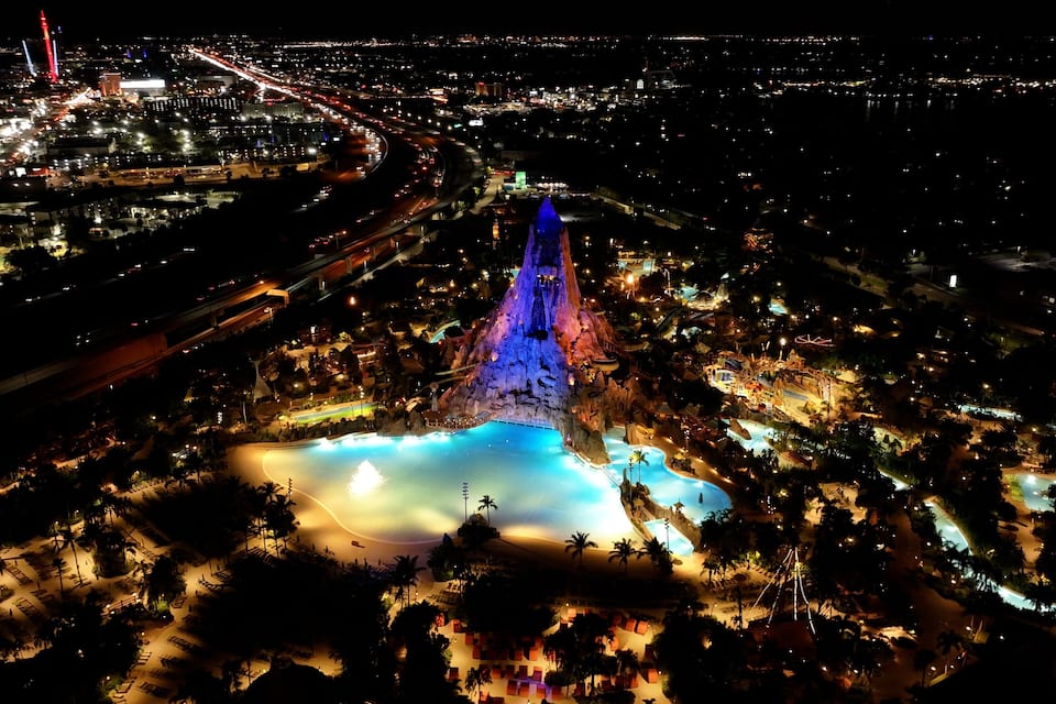 Vue aérienne d’une ville de nuit, avec une montagne illuminée qui se détache sur le paysage et une grande zone de piscine.