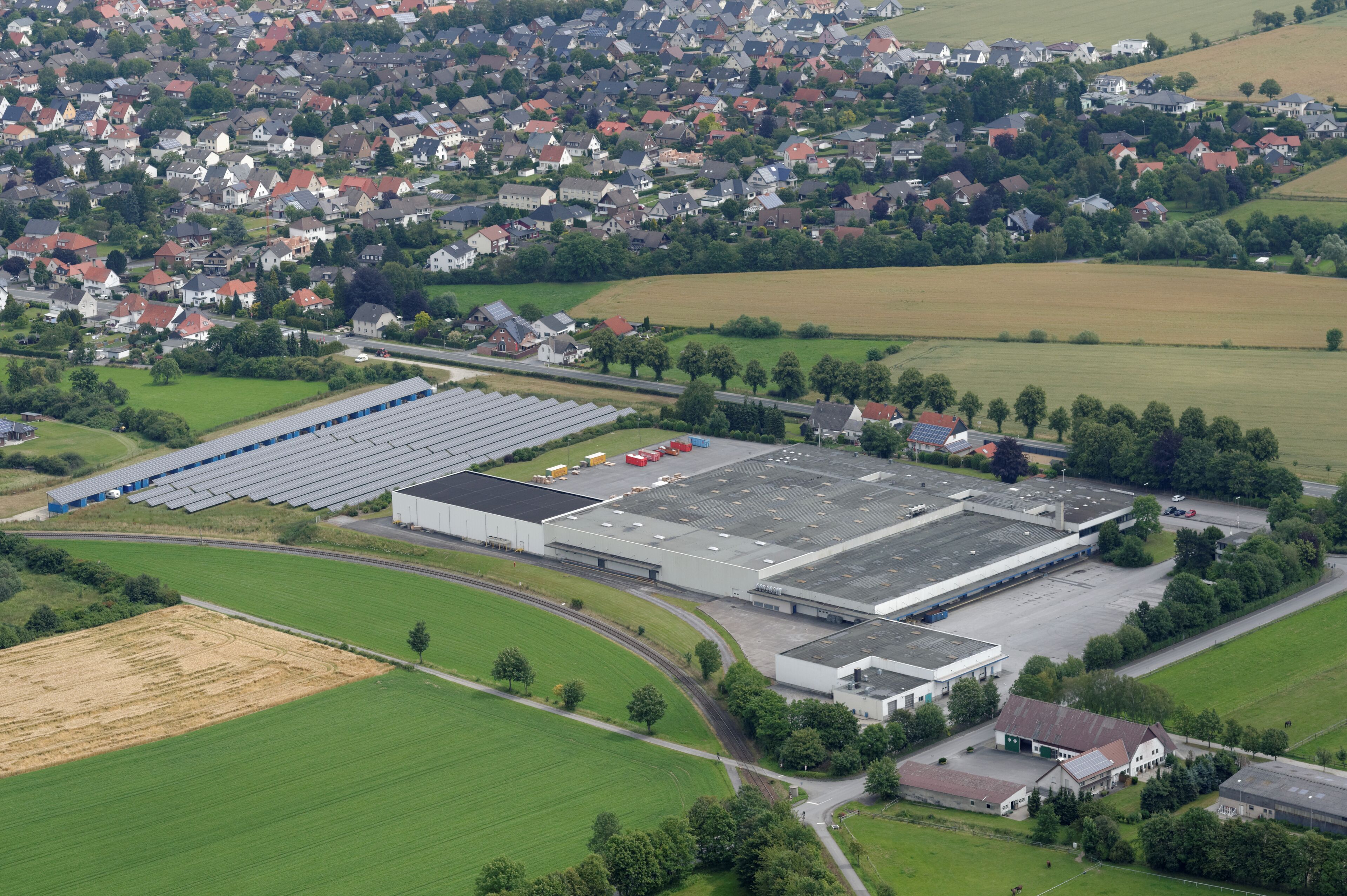 An industrial area with large buildings, a parking lot, and surrounding green fields and residential houses.