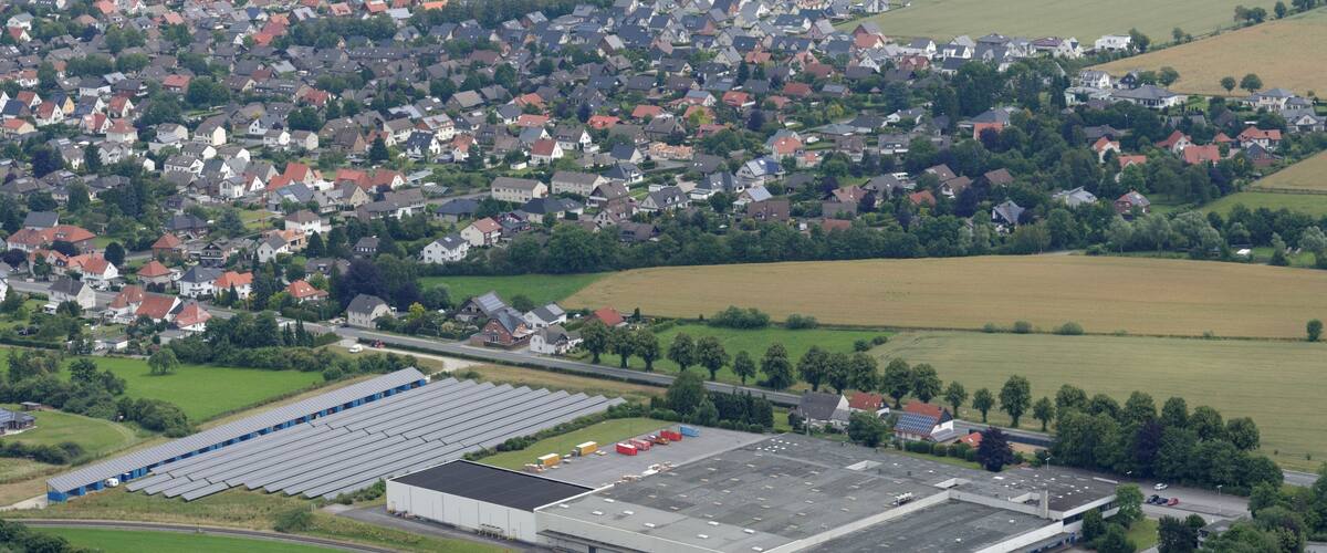 An industrial area with large buildings, a parking lot, and surrounding green fields and residential houses.
