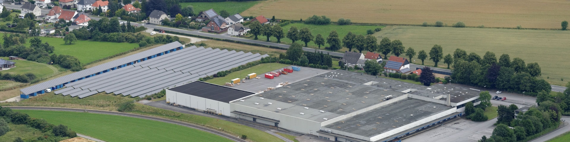 An industrial area with large buildings, a parking lot, and surrounding green fields and residential houses.