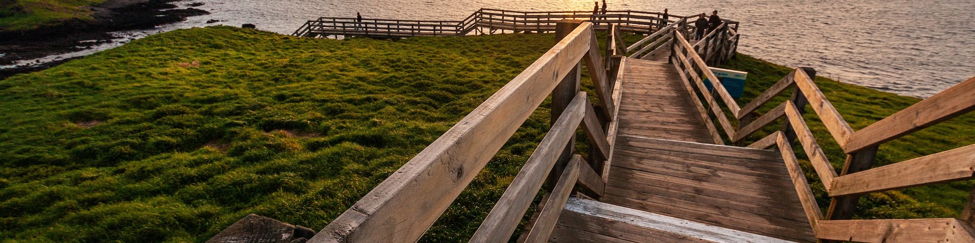 A wooden boardwalk leading to a grassy area with a view of the ocean and a distant island at sunset.