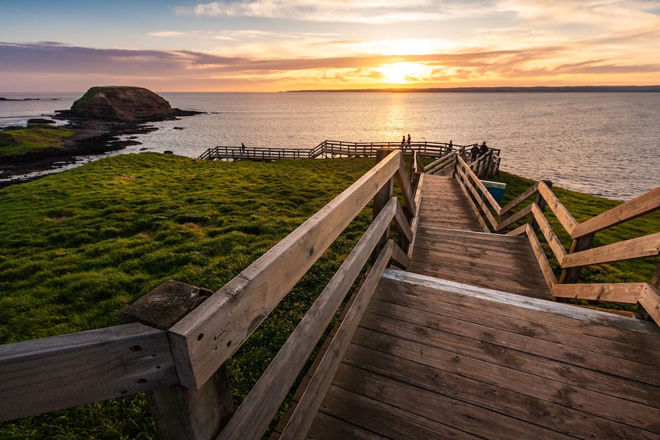 A wooden boardwalk leading to a grassy area with a view of the ocean and a distant island at sunset.