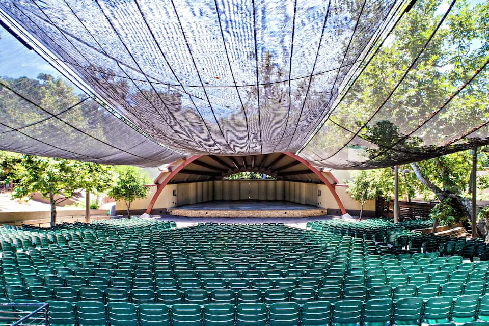 Een groot openluchttheater met groene zitplaatsen en een podium onder een overkapping.