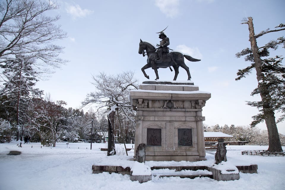 A snow-covered statue of a mounted figure on a pedestal in a park with trees and a building in the background.