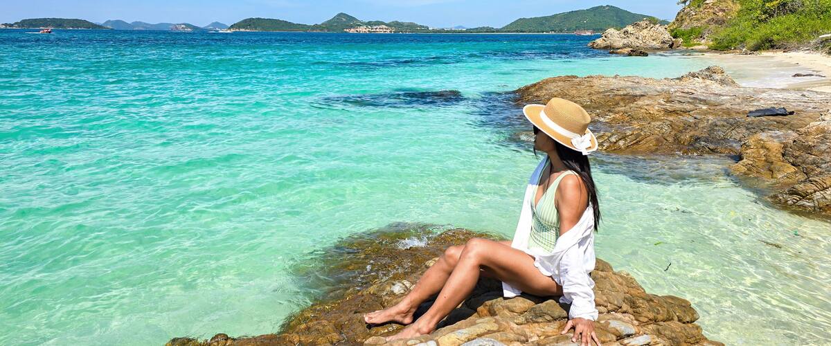 A woman sitting on rocks by a turquoise sea with clear water, surrounded by greenery and distant hills.