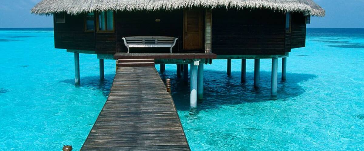 A wooden overwater bungalow with a thatched roof, connected to the shore by a wooden pier.