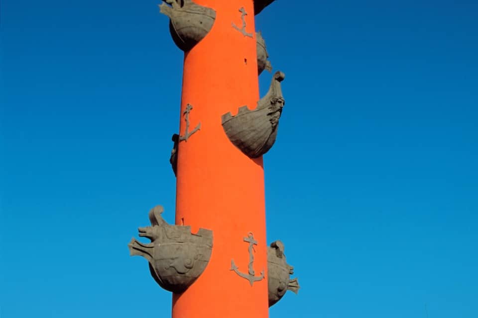 Un'alta colonna arancione con statue raffiguranti una donna e una barca, circondata da edifici.