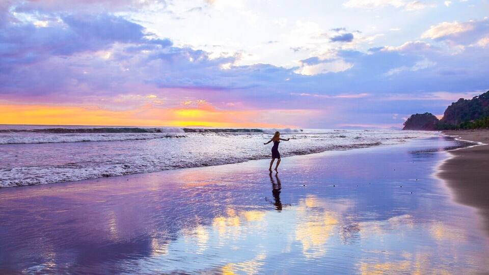 A beach at sunset with waves, a person, and a rocky cliff.
