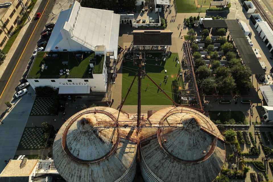 Una vista aerea di un ampio complesso industriale con due silos ben visibili, un'area di parcheggio e un campo verde.