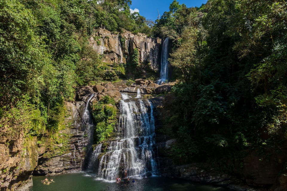 A waterfall cascading into a pool surrounded by dense forest and rocky cliffs.